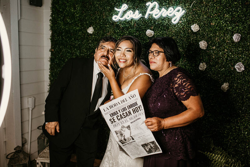 bride with parents posing in front of greenery wall with neon sign photo booth