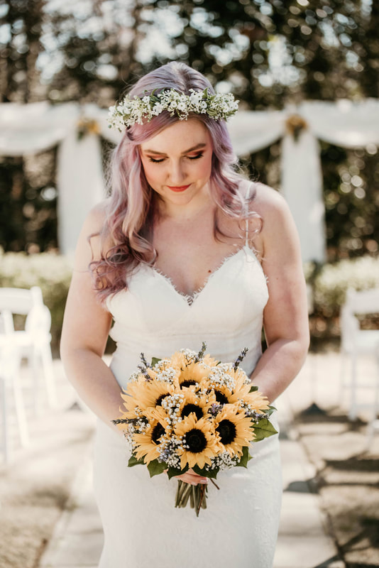 bride posing by outdoor ceremony area looking down at bouquet of sunflowers