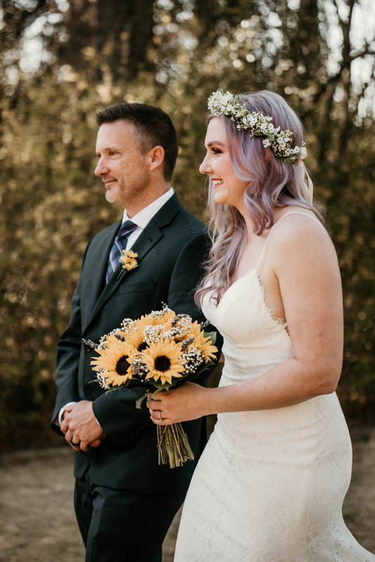 bride with lilac hair wearing a flower crown holding sunflower bouquet walking with dad