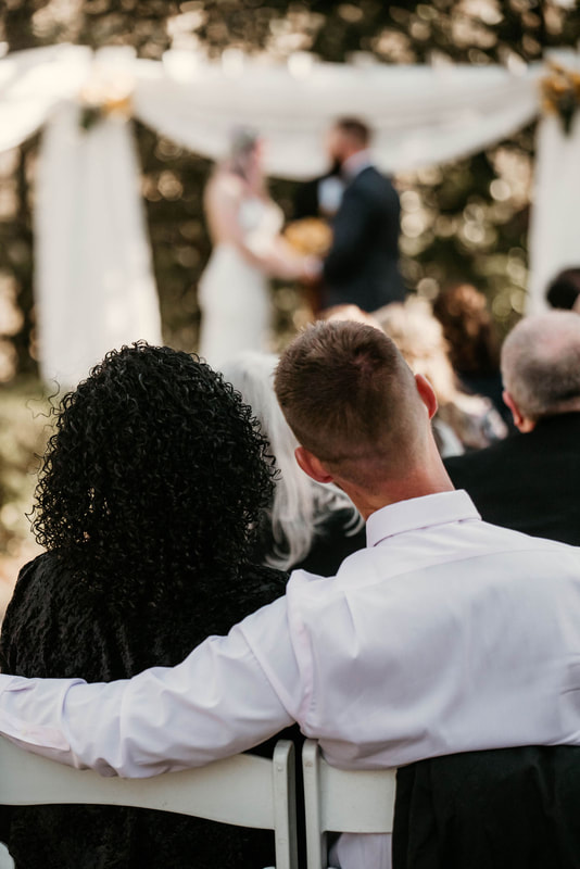 man with arm around woman's shoulder as they watch wedding ceremony