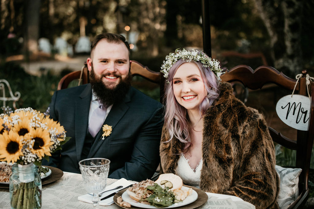 couple at sweetheart table. bride wearing faux fur coat over dress with flower crown in her hair